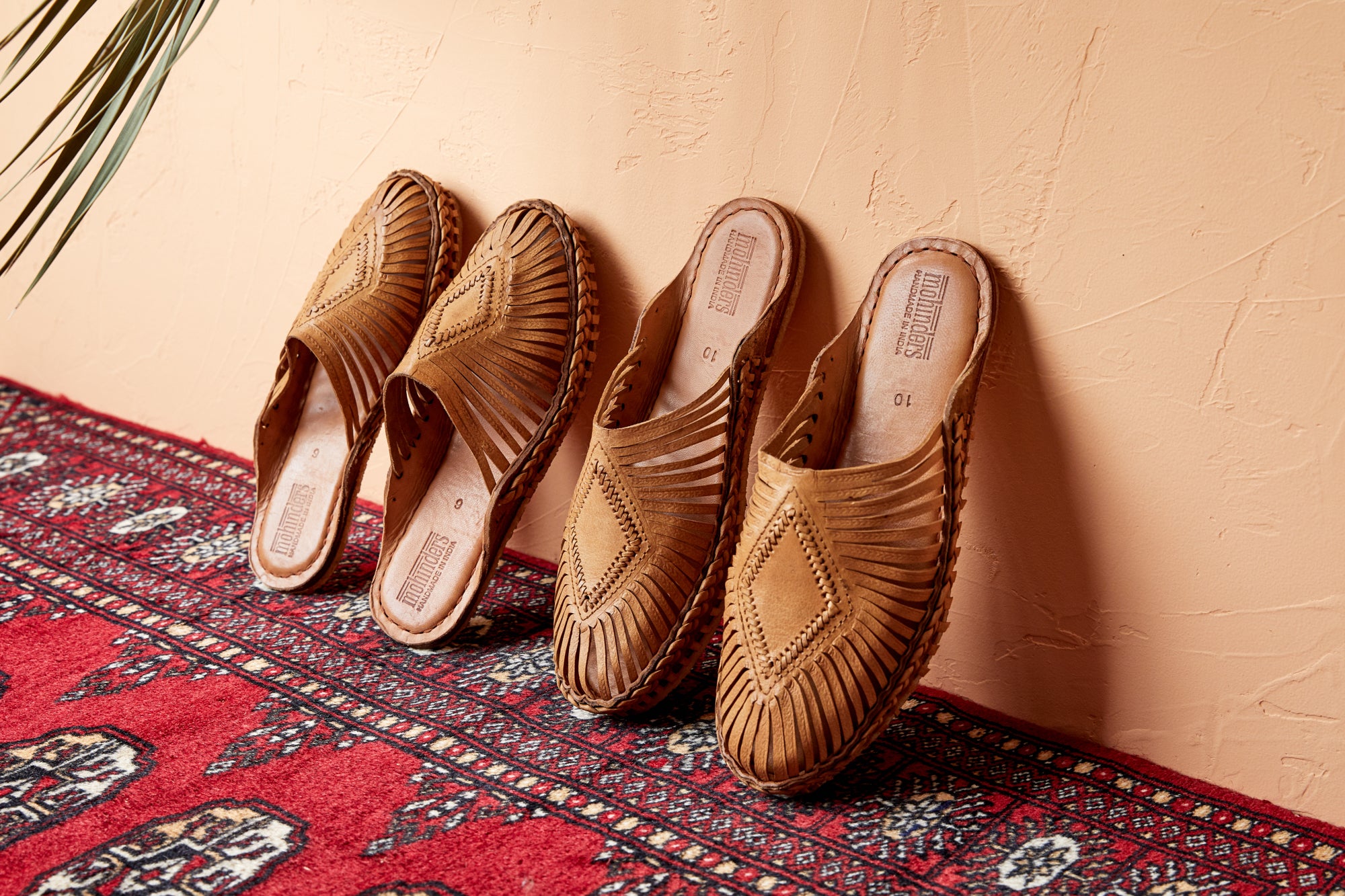 Two pairs of natural leather slip-on sandals with hand-woven diamond centerpiece on red rug propped against wall